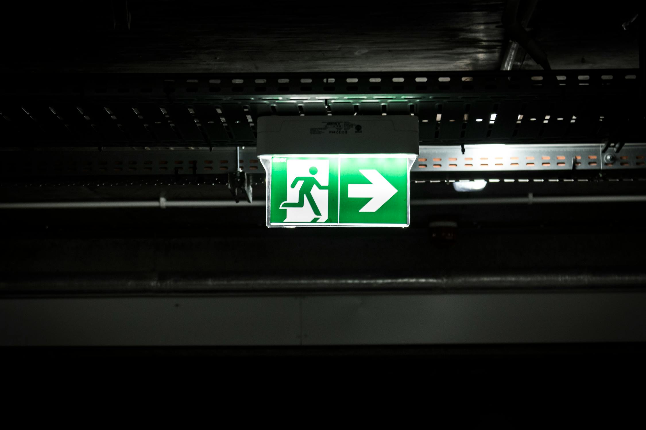 Illuminated green emergency exit sign with running man pictogram on dark industrial ceiling